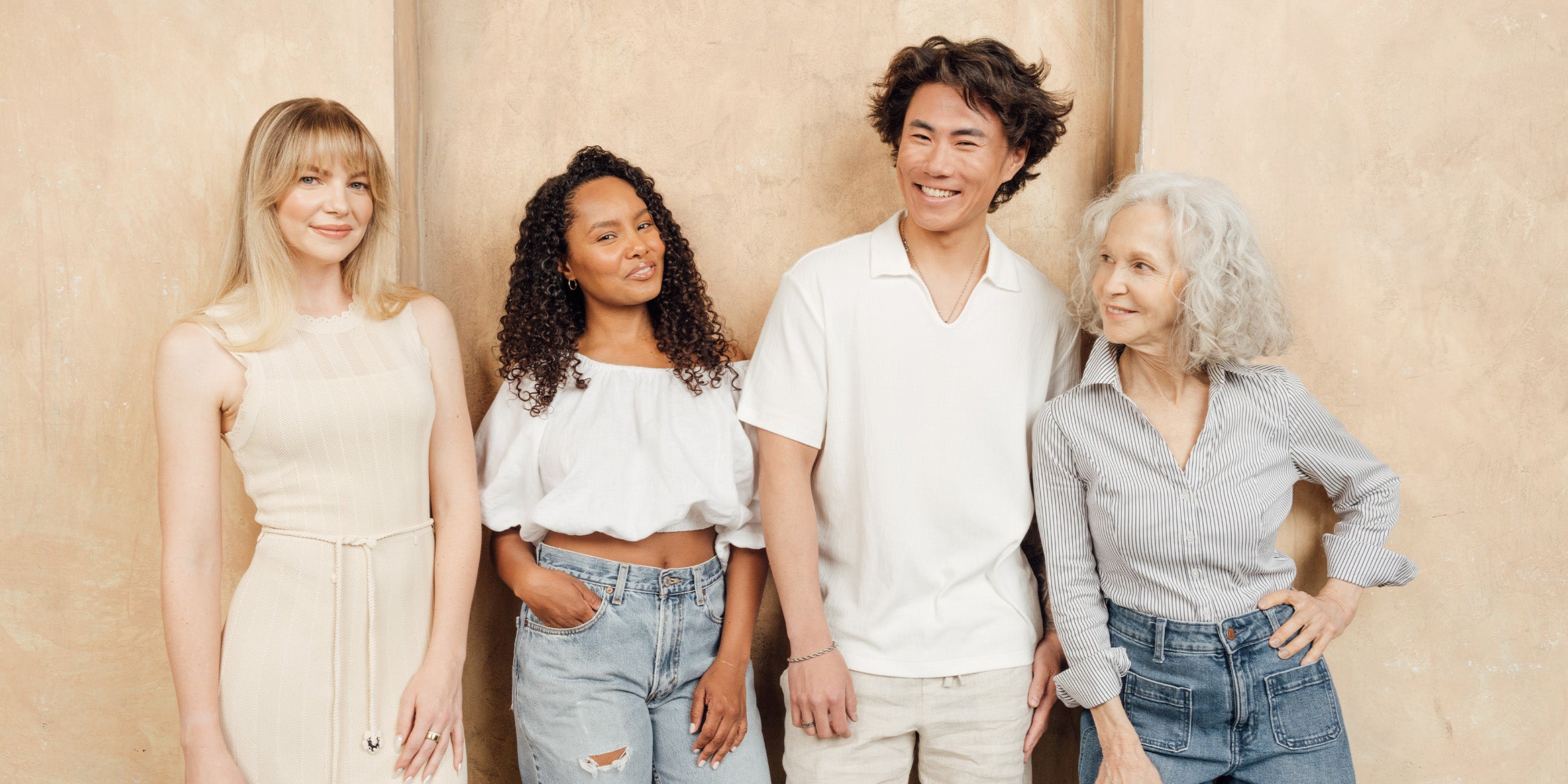 a young blonde white woman, a young black woman, a young asian man, and a senior white woman, all in a group standing next to each other and posing for the camera against a tan color wall
