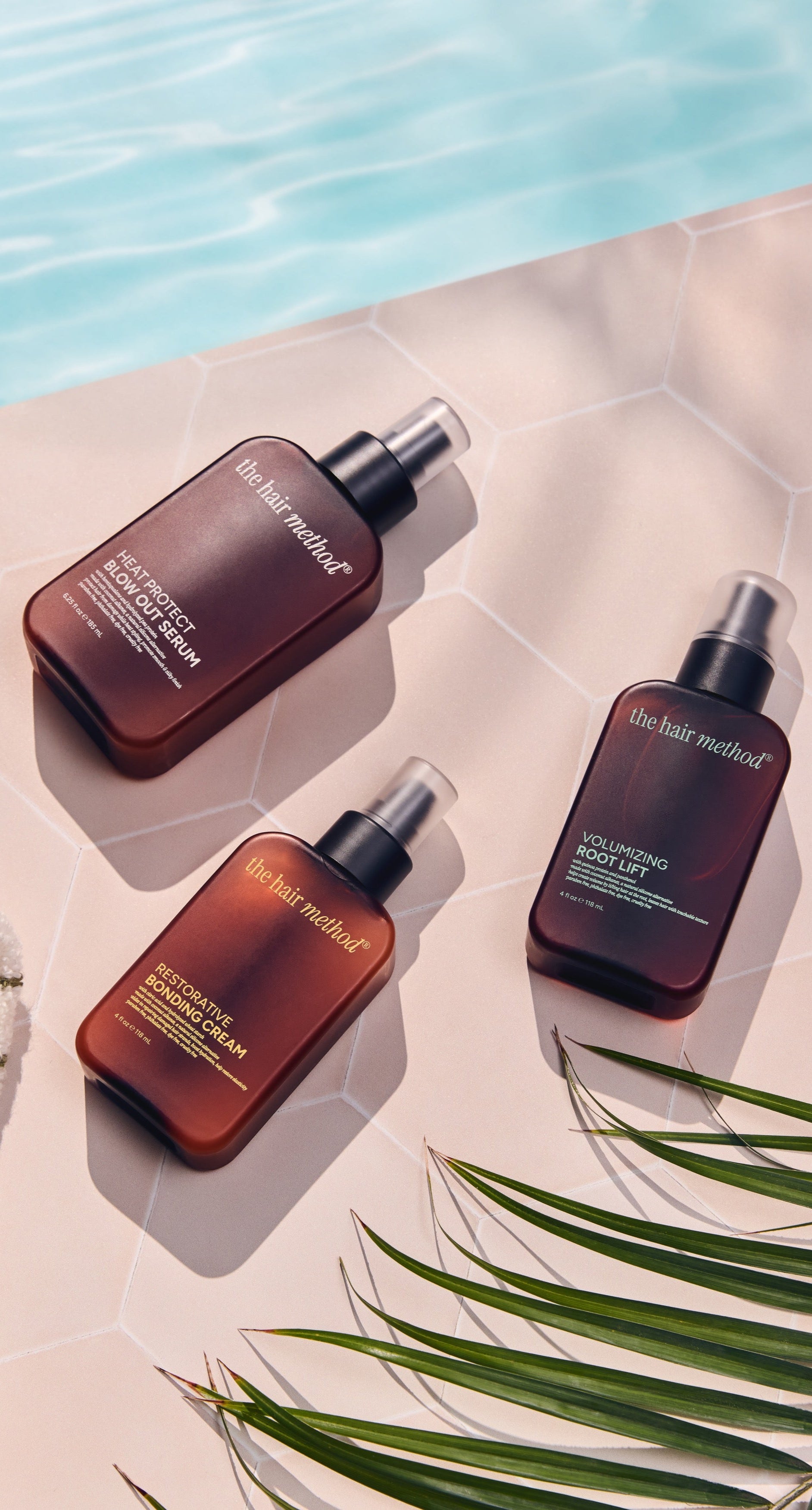 Three bottles of 'The Hair Method' product on a poolside surface with a towel and palm leaf.