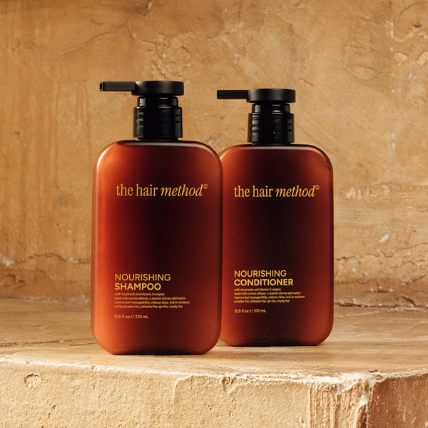 photograph of two brown bottles with black pumps and light text printed on them. the products are on a yellow step with a yellow weathered wall in the background. The bottles read The Hair Method Nourishing Shampoo and Conditioner, from left to right respectively.