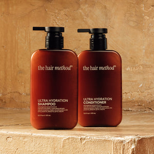 photograph of two brown bottles with black pumps and light text printed on them. the products are on a yellow step with a yellow weathered wall in the background. The bottles read The Hair Method Ultra Hydration Shampoo and Conditioner, from left to right respectively.