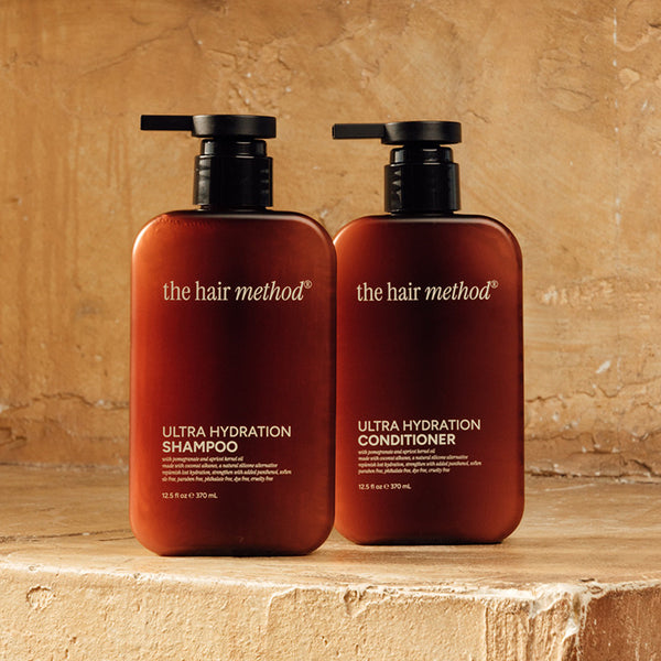 photograph of two brown bottles with black pumps and light text printed on them. the products are on a yellow step with a yellow weathered wall in the background. The bottles read The Hair Method Ultra Hydration Shampoo and Conditioner, from left to right respectively.