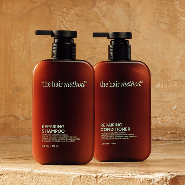 photograph of two brown bottles with black pumps and light text printed on them. the products are on a yellow step with a yellow weathered wall in the background. The bottles read The Hair Method Repairing Shampoo and Conditioner, from left to right respectively.