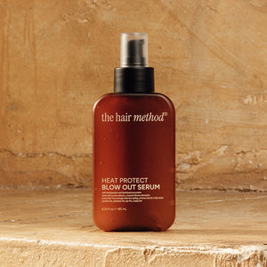 photograph of the front of a brown bottle with black pump and light gray text printed on it. the product is on a yellow weathered background with a textured wall behind it. it reads The Hair Method Heat Protect Blow Out Serum.