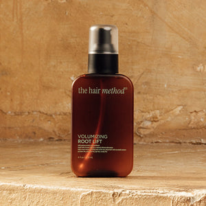 photograph of the front of a brown bottle with black pump and light green text printed on it. the product is on a yellow weathered background with a textured wall behind it. it reads The Hair Method Volumizing Root Lift.