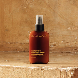 photograph of the front of a brown bottle with black pump and light orange text printed on it. the product is on a yellow weathered background with a textured wall behind it. it reads The Hair Method Restorative Bonding Cream.