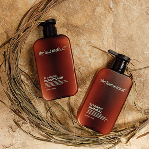 A photo of a shampoo and conditioner set in brown bottles laying flat against a textured tan surface with dried palm leaves in a half circle around them