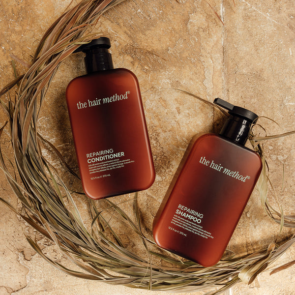 A photo of a shampoo and conditioner set in brown bottles laying flat against a textured tan surface with dried palm leaves in a half circle around them