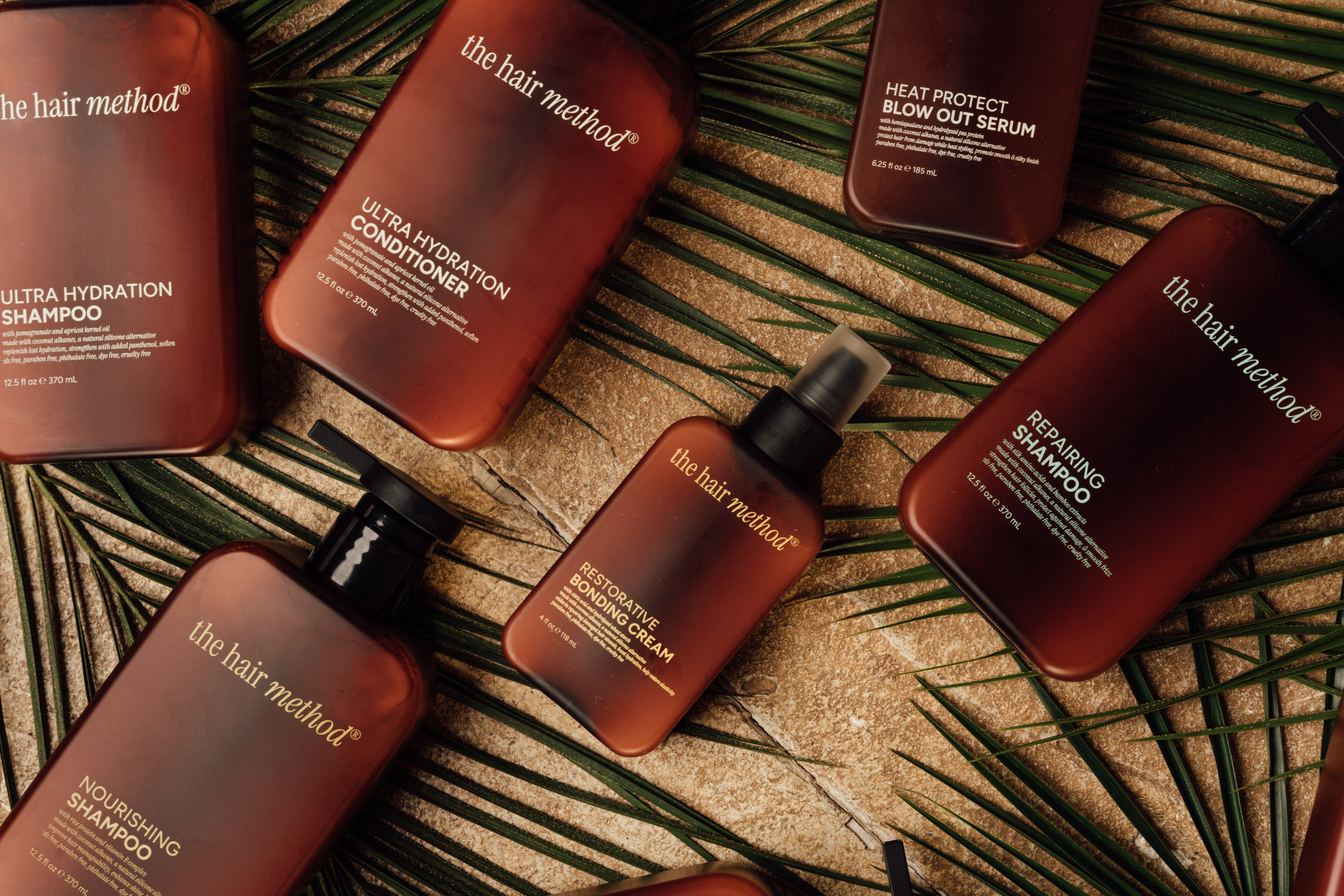 A photo of multiple beauty products laid on a tan surface with a palm leaf underneath them