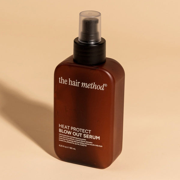 photograph of an angled brown bottle with black pump and light gray text printed on it. the product is on a tan background with a large shadow behind it. it reads The Hair Method Heat Protect Blow Out Serum.