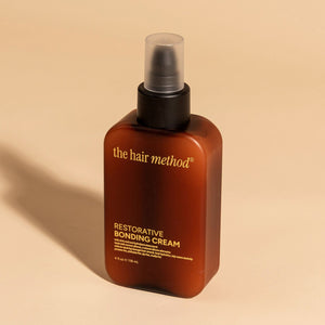 photograph of an angled brown bottle with black pump and light orange text printed on it. the product is on a tan background with a large shadow behind it. it reads The Hair Method Restorative Bonding Cream.