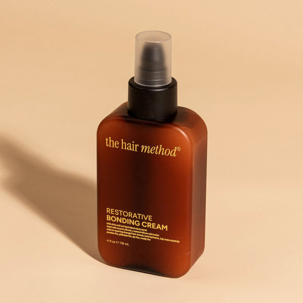 photograph of an angled brown bottle with black pump and light orange text printed on it. the product is on a tan background with a large shadow behind it. it reads The Hair Method Restorative Bonding Cream.
