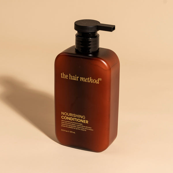 photograph of an angled brown bottle with black pump and light orange text printed on it. the product is on a tan background with a large shadow behind it. it reads The Hair Method Nourishing Conditioner.
