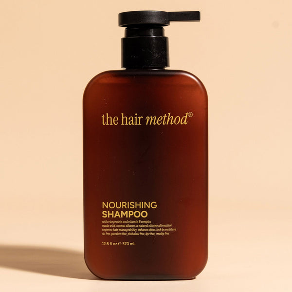 photograph of the front of a brown bottle with black pump and light gray text printed on it. the product is on a tan background with a large shadow behind it. it reads The Hair Method Nourishing Shampoo.