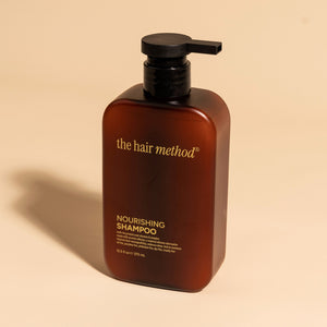 photograph of an angled brown bottle with black pump and light orange text printed on it. the product is on a tan background with a large shadow behind it. it reads The Hair Method Nourishing Shampoo.