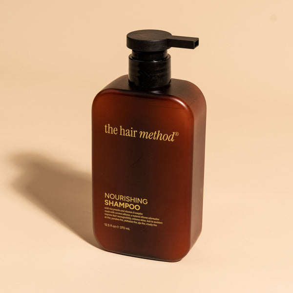 photograph of an angled brown bottle with black pump and light orange text printed on it. the product is on a tan background with a large shadow behind it. it reads The Hair Method Nourishing Shampoo.