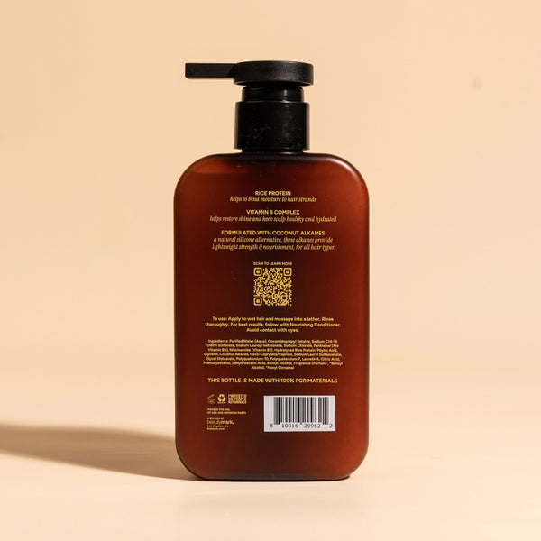 photograph of the back of a brown bottle with black pump and light green text printed on it. the product is on a tan background with a large shadow behind it