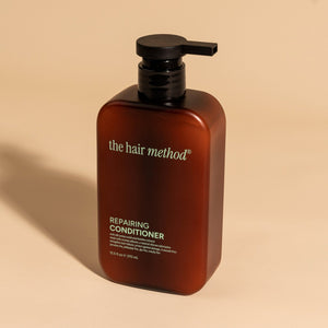 photograph of an angled brown bottle with black pump and light green text printed on it. the product is on a tan background with a large shadow behind it. it reads The Hair Method Repairing Conditioner.