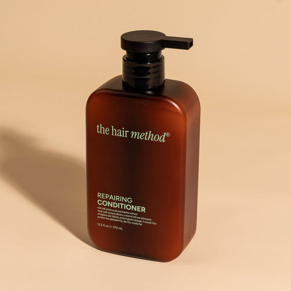 photograph of an angled brown bottle with black pump and light green text printed on it. the product is on a tan background with a large shadow behind it. it reads The Hair Method Repairing Conditioner.