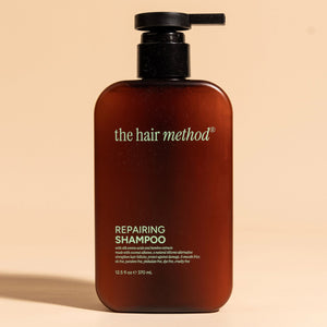 photograph of the front of a brown bottle with black pump and light gray text printed on it. the product is on a tan background with a large shadow behind it. it reads The Hair Method Repairing Shampoo.