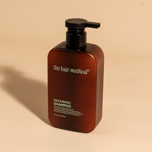 photograph of an angled brown bottle with black pump and light green text printed on it. the product is on a tan background with a large shadow behind it. it reads The Hair Method Repairing Shampoo.