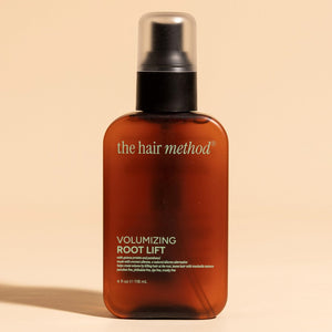 photograph of the front of a brown bottle with black pump and light green text printed on it. the product is on a tan background with a large shadow behind it. it reads The Hair Method Volumizing Root Lift.