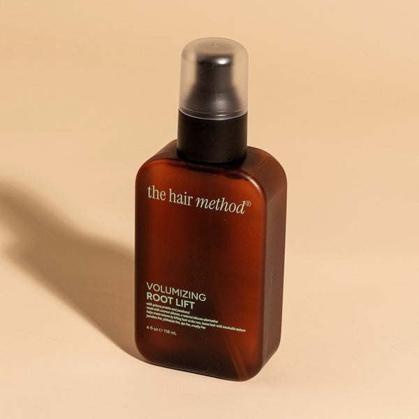 photograph of an angled brown bottle with black pump and light green text printed on it. the product is on a tan background with a large shadow behind it. it reads The Hair Method Volumizing Root Lift.