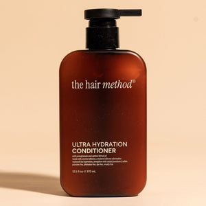 photograph of the front of a brown bottle with black pump and light gray text printed on it. the product is on a tan background with a large shadow behind it. it reads The Hair Method Ultra Hydration Conditioner.