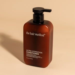 photograph of an angled brown bottle with black pump and light gray text printed on it. the product is on a tan background with a large shadow behind it. it reads The Hair Method Ultra Hydration Conditioner.
