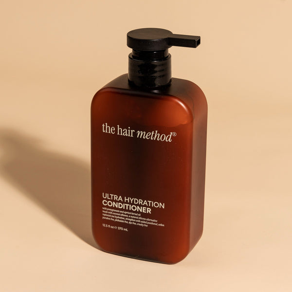 photograph of an angled brown bottle with black pump and light gray text printed on it. the product is on a tan background with a large shadow behind it. it reads The Hair Method Ultra Hydration Conditioner.