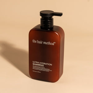 photograph of an angled brown bottle with black pump and light gray text printed on it. the product is on a tan background with a large shadow behind it. it reads The Hair Method Ultra Hydration Shampoo.