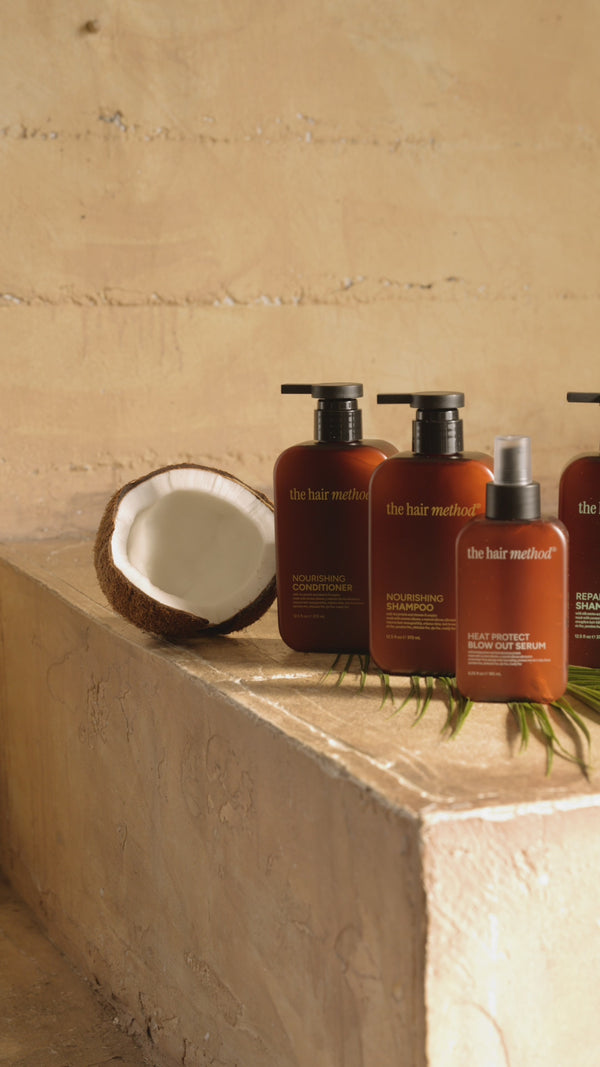 A slow panning video of haircare products lined up on a yellow tan step with a textured wall behind them. There is a cut coconut and palm leaves used as props, with natural light hitting all products in their brown bottles. There is no audio.