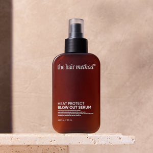 Bottle of 'the hair method' heat protect blowout serum on a beige surface with a neutral background