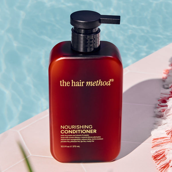 Amber bottle of 'the hair method' conditioner by a poolside with a towel and plant.