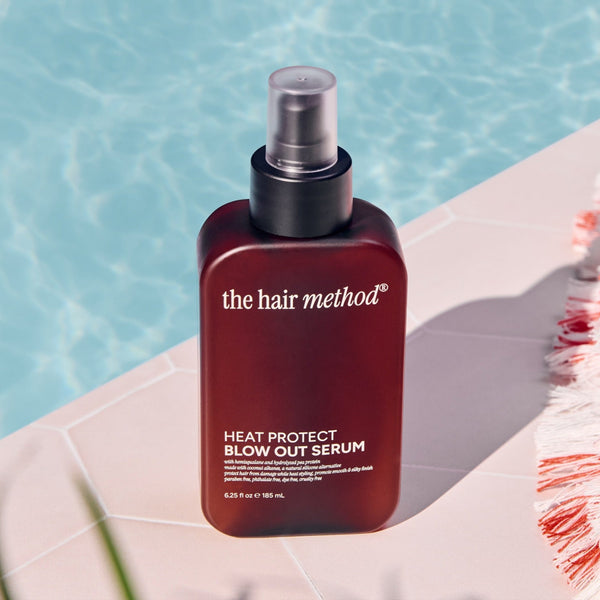 Amber bottle of 'the hair method' heat protect blowout serum on a white surface with a blurred blue background