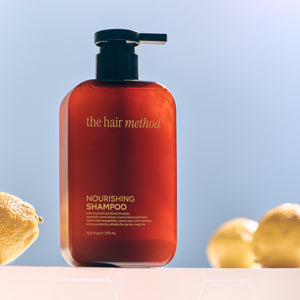Amber shampoo bottle with pump labeled 'the hair method' on a light blue background with lemons and leaves.