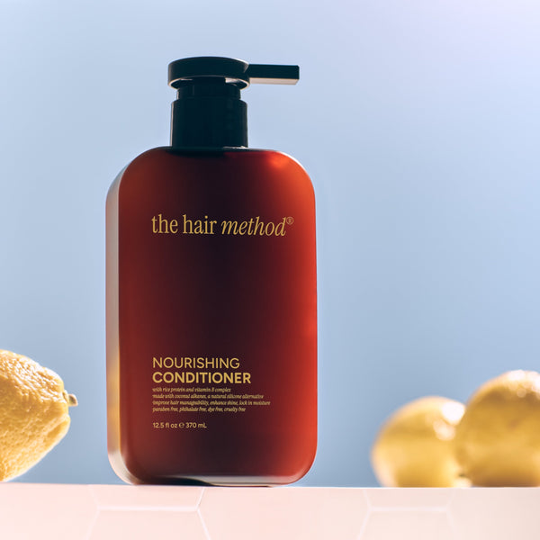 Amber bottle of 'the hair method' nourishing conditioner with lemons and leaves on a light blue background